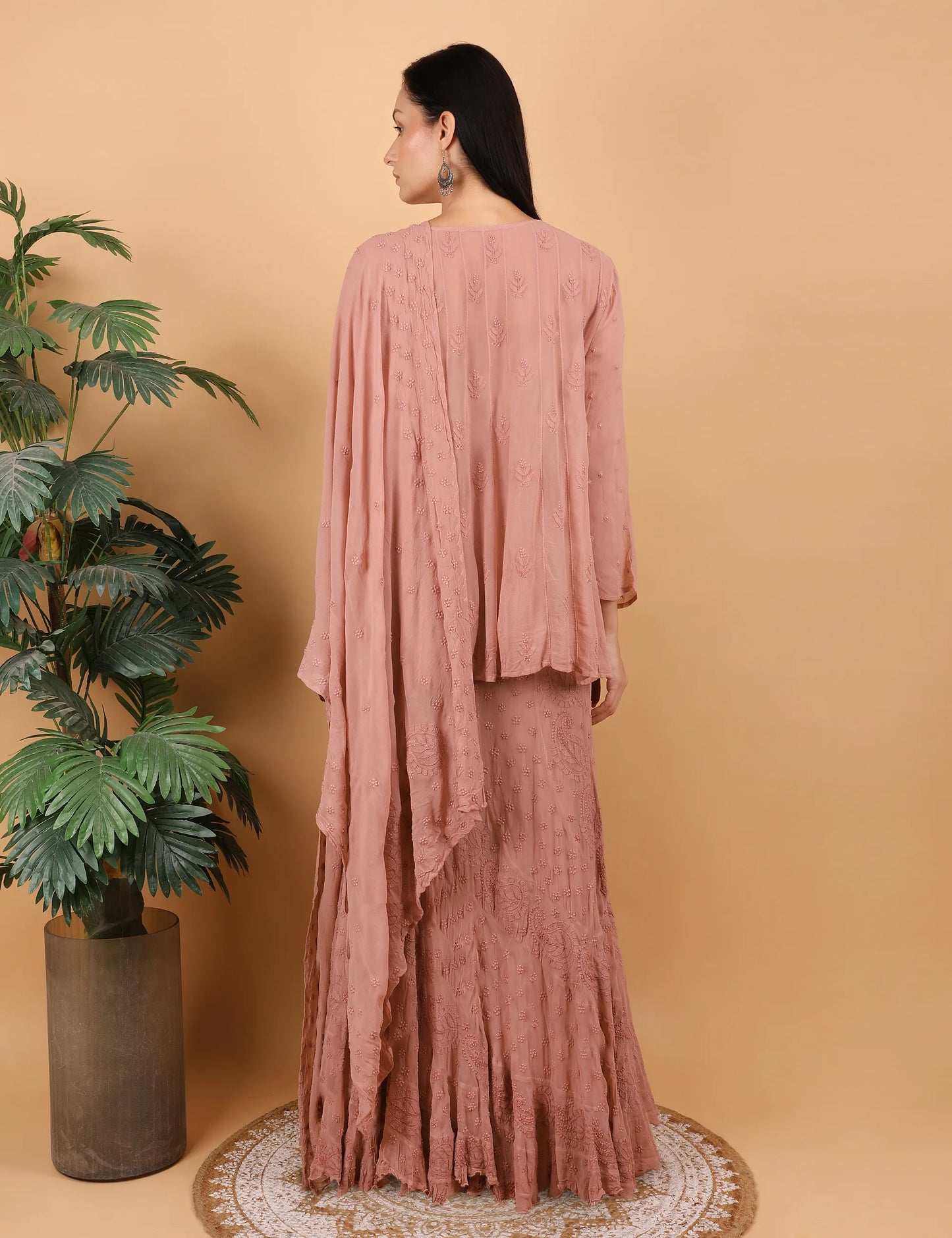 Woman wearing a Hand Embroidered chikankari lahenga and choli Peach against a beige wall with a plant to her left.