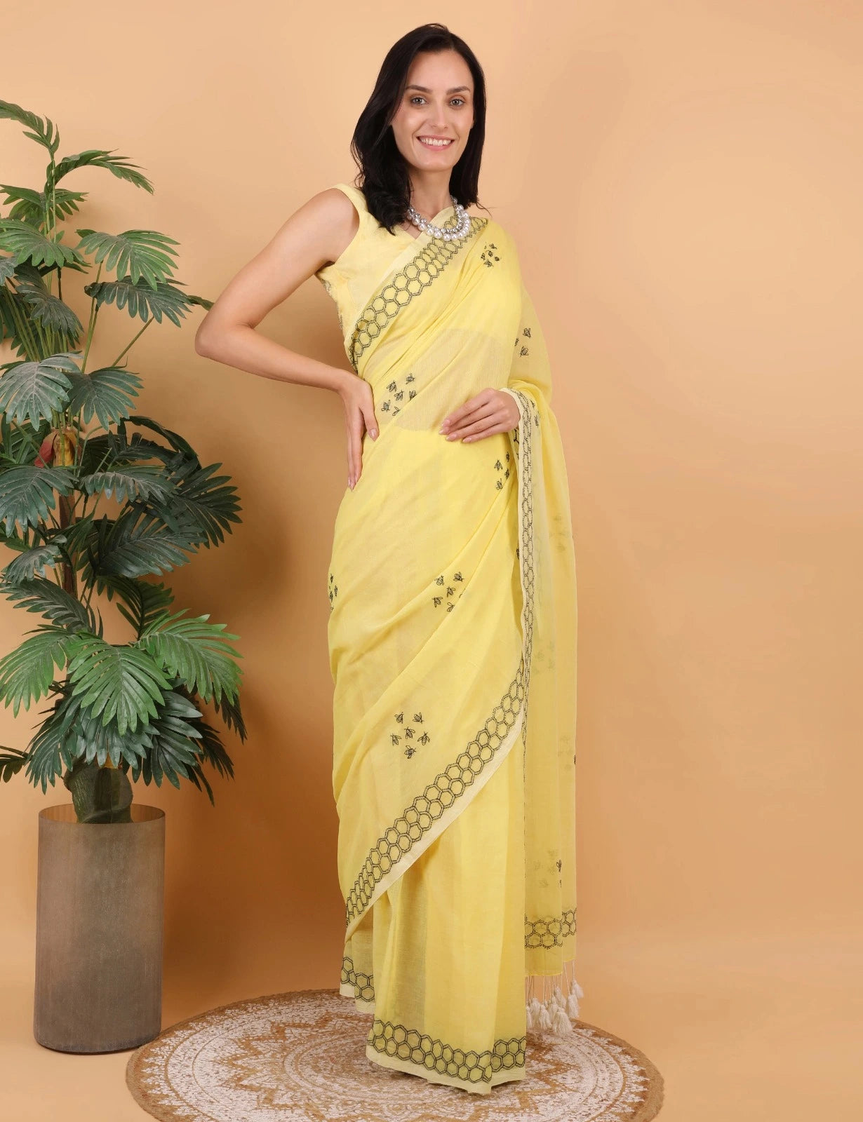 Woman in a yellow Mulmul saree standing next to a potted plant on a beige background