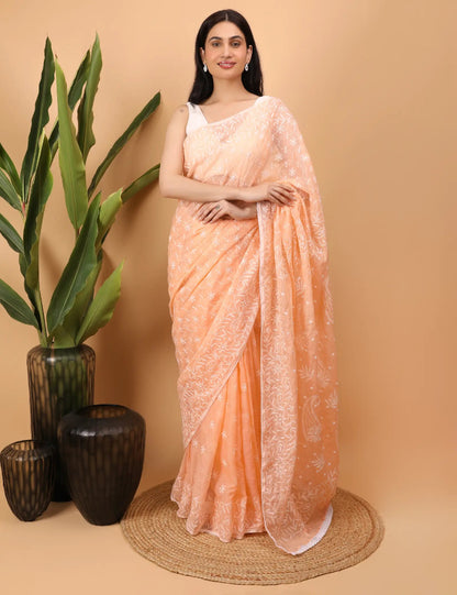 Woman in a peach Hand Embroidered  Chikankari saree standing against a beige background with decorative vases and plants.