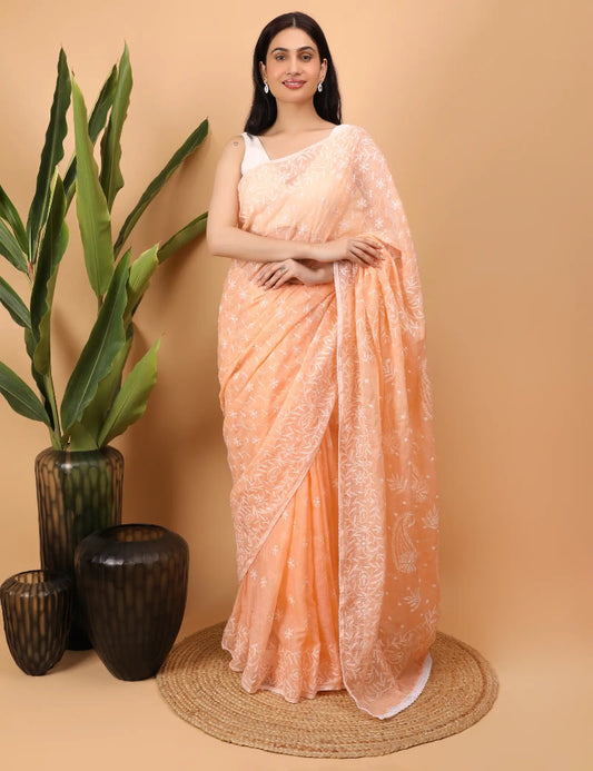 Woman in a peach Hand Embroidered  Chikankari saree standing against a beige background with decorative vases and plants.