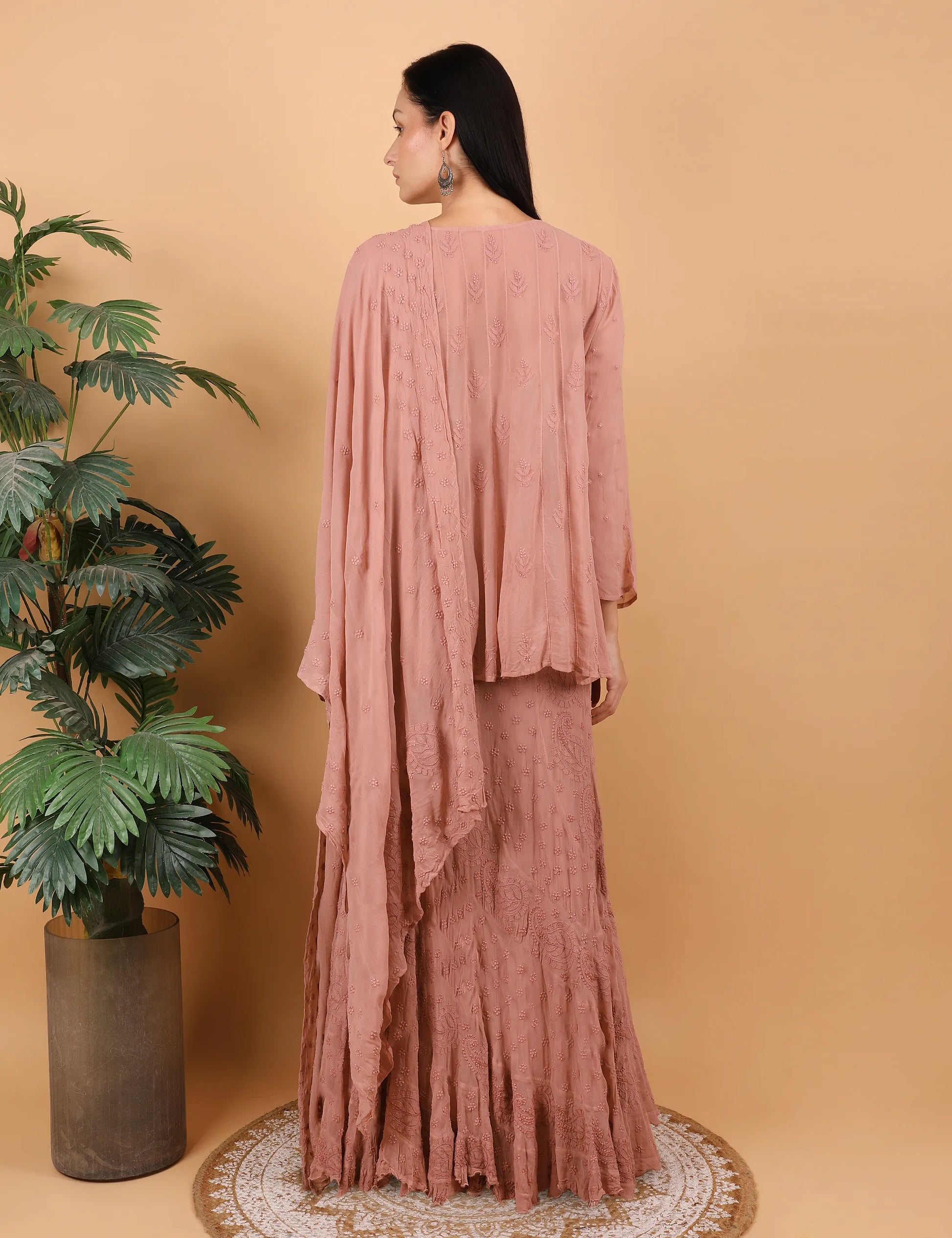 Woman wearing a  Hand Embroidered chikankari lahenga and choli Peach against a beige wall with a plant to her left.