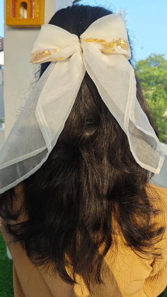 Person wearing a large white bow with gold accents on their head outdoors.