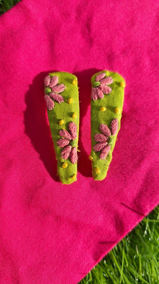 Two green hair clips with pink Hand embroidered chikankari designs on a pink fabric background.
