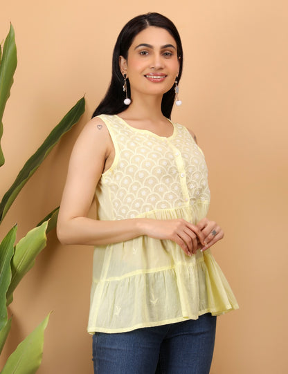 Shwet poses with a bright smile, wearing a sleeveless lemon yellow peplum-style cotton short top with delicate white Chikankari embroidery. The top features a scallop-like floral pattern on the upper bodice and tiered panels with subtle motifs. She pairs it with dark blue jeans and accessorizes with statement earrings. Green leaves frame the composition against a soft beige background.