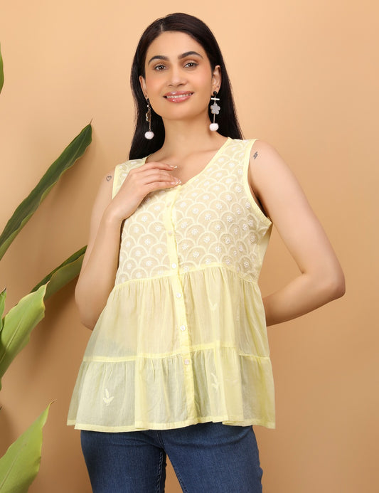 Shwet smiles softly in a close-up , wearing a sleeveless lemon yellow top adorned with delicate white Chikankari embroidery in a scalloped floral pattern. She accessorizes with statement drop earrings featuring pom-poms and floral elements. Her hand gently touches her neck, showcasing a small lightning bolt tattoo on her upper arm. A warm beige background and green leaves add a fresh, natural touch to the composition.









