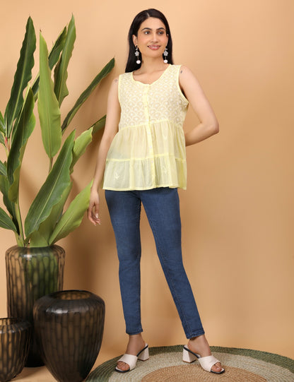 Shwet smiles softly in wearing a sleeveless lemon yellow top adorned with delicate white Chikankari embroidery in a scalloped floral pattern. She accessorizes with statement drop earrings featuring pom-poms and floral elements,showcasing a small lightning bolt tattoo on her upper arm. A warm beige background and green leaves add a fresh, natural touch to the composition. Yellow Chikankari Tier top paired with jeans . Styled with white block heels. 