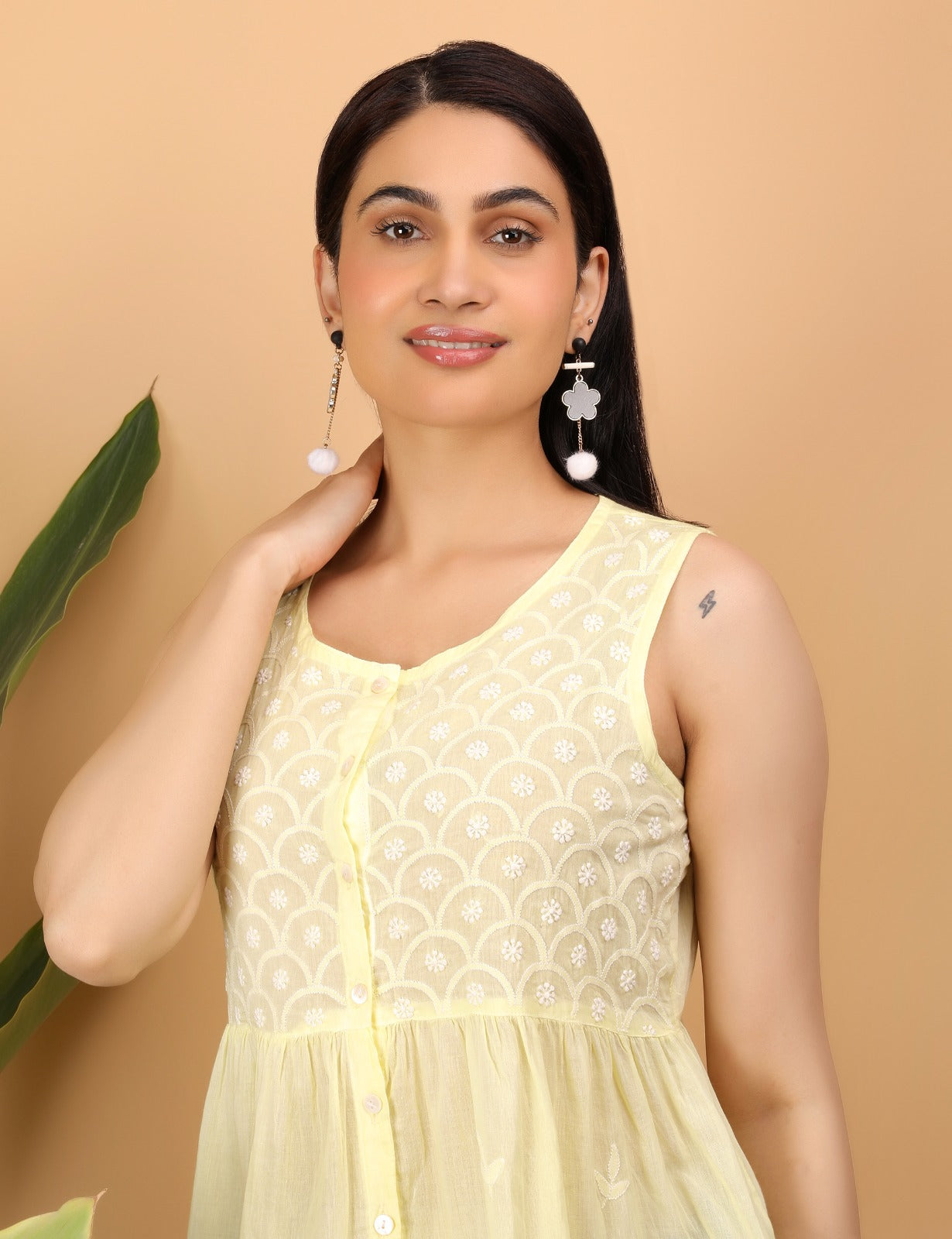 Shwet smiles softly in a close-up portrait, wearing a sleeveless lemon yellow top adorned with delicate white Chikankari embroidery in a scalloped floral pattern. She accessorizes with statement drop earrings featuring pom-poms and floral elements. Her hand gently touches her neck, showcasing a small lightning bolt tattoo on her upper arm. A warm beige background and green leaves add a fresh, natural touch to the composition.