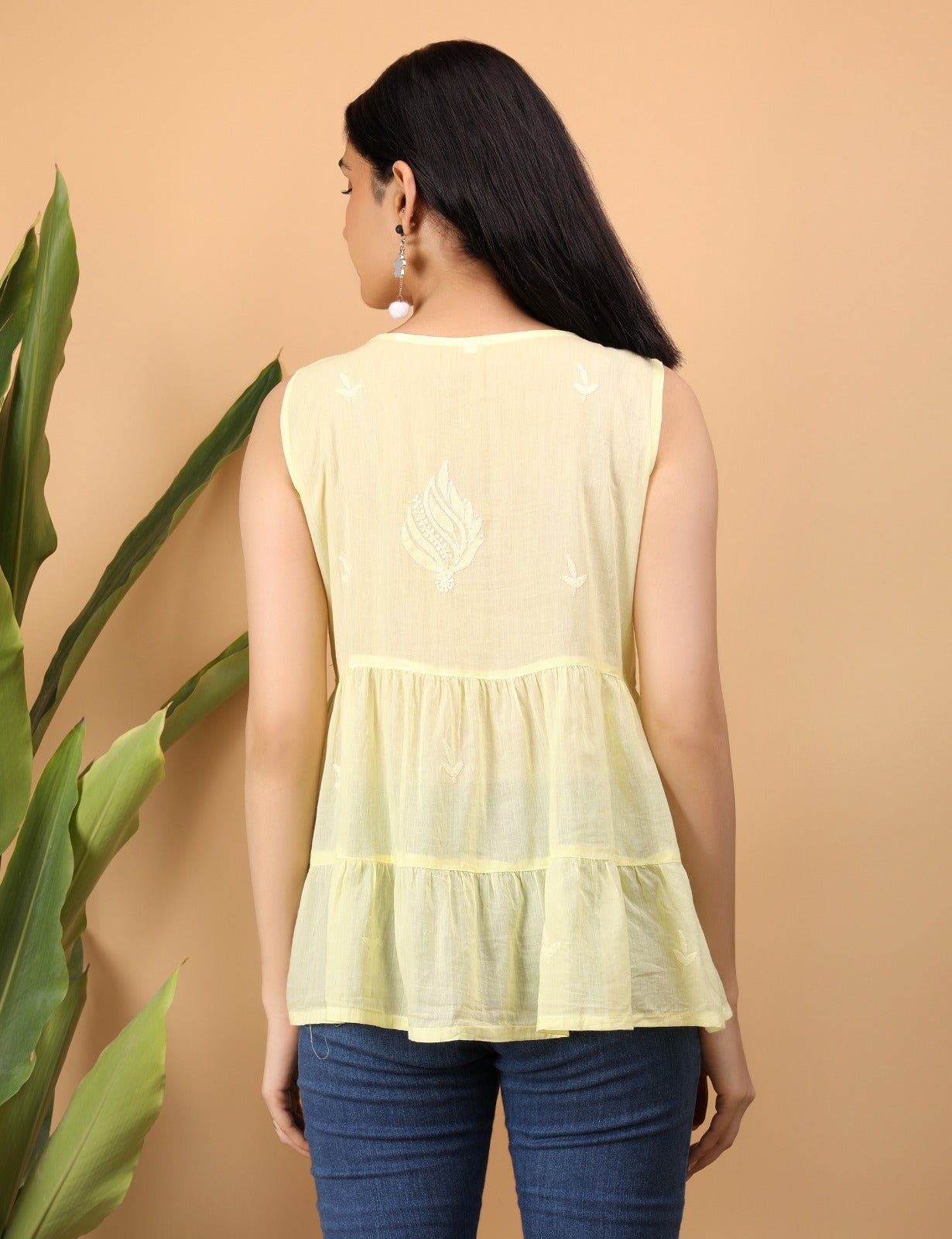 Shwet is seen from the back, wearing a sleeveless lemon yellow cotton top featuring delicate white Chikankari embroidery. The top has a flared, tiered design with a central embroidered paisley motif and scattered floral details. Her long black hair falls straight down, and she accessorizes with dangling earrings. She stands against a warm beige background with green leaves partially visible on the side.









